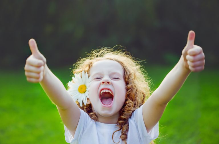 A little girl with a flower in her curly red hair holding two thumbs up enthusiastically while standing in a field.